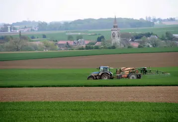 Tracteur fertilisant un champ devant un village 