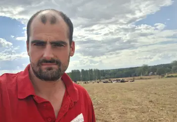 Un jeune éleveur devant sa prairie jaunie avec ses génisses au fond